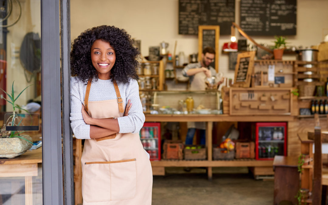 Portrait of a smiling young African American barista leaning with her arms crossed on the door of a trendy cafe